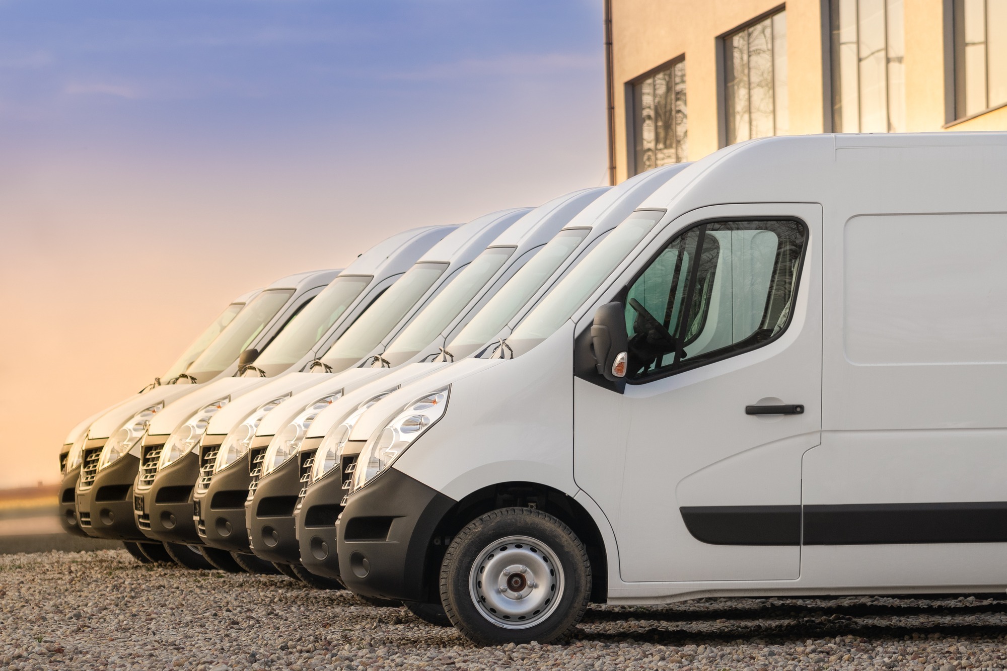 A row of identical white delivery vans parked on a gravel lot, showcasing their streamlined design against a twilight sky.