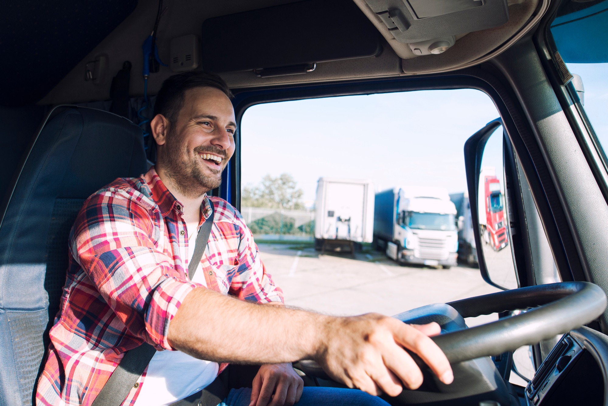 Truck driver smiling while steering, showcasing a positive attitude in a commercial vehicle park. Ideal for illustrating the trucking lifestyle.